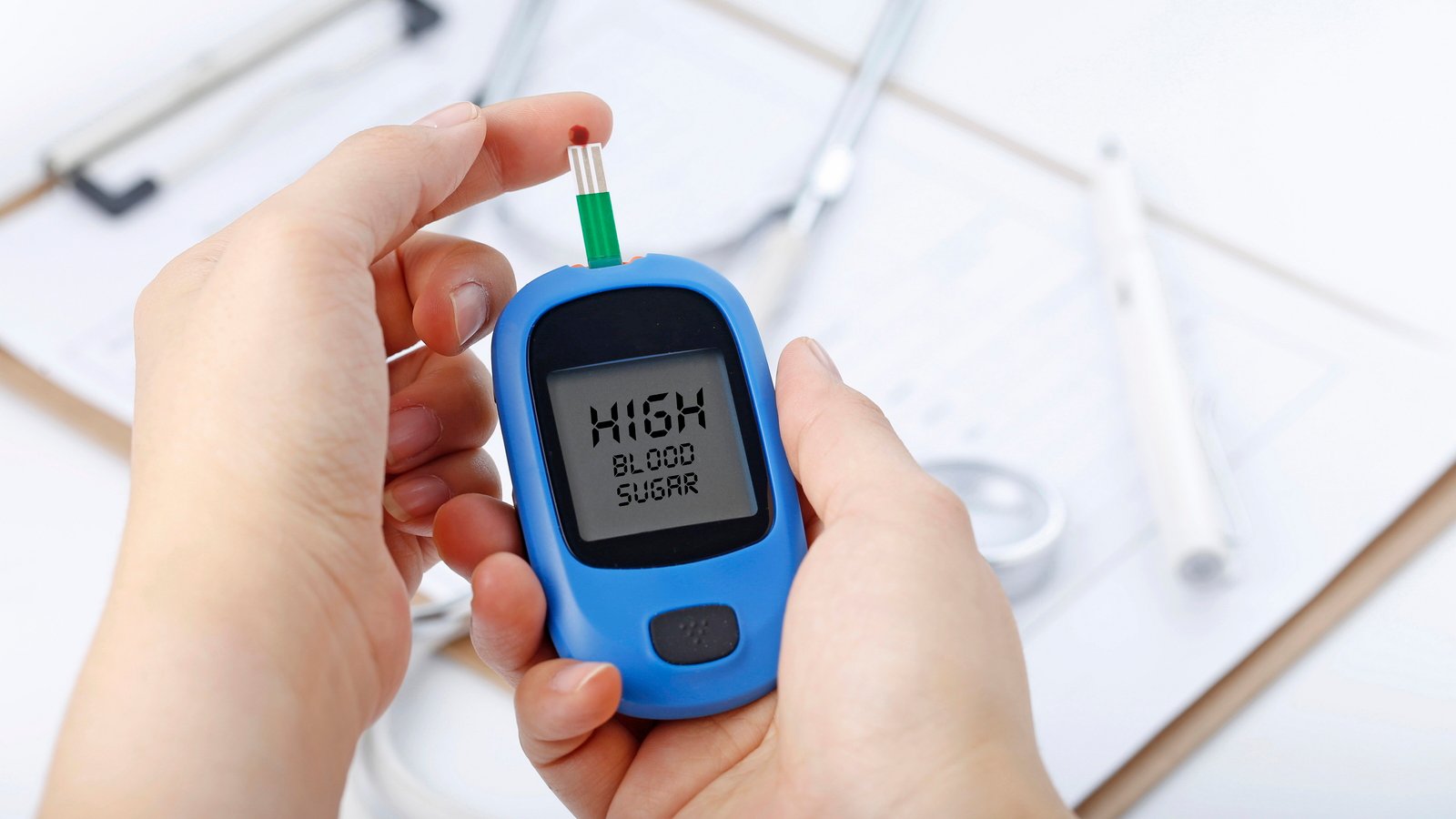 Hand holding a blood glucose meter measuring blood sugar, the background is a stethoscope and chart file